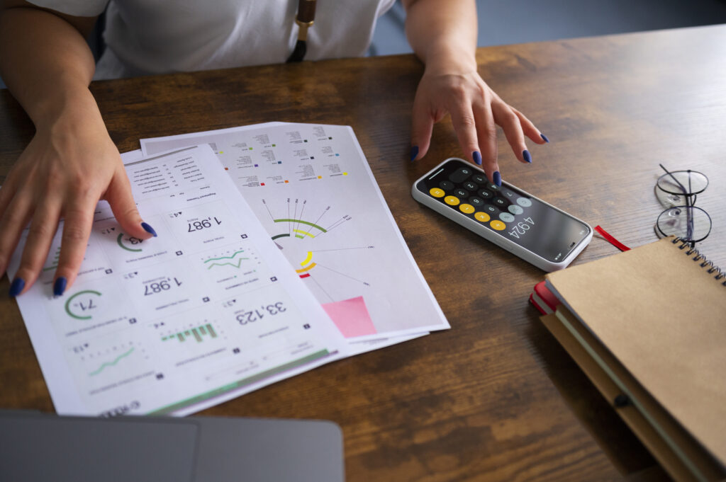 A person reviewing financial reports and charts on paper while using a smartphone calculator, symbolizing efficient business management and decision-making.