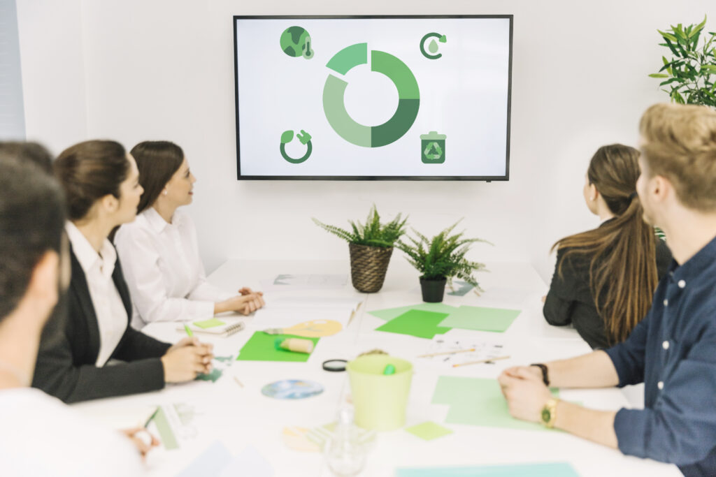 A team of professionals in a meeting room discussing a presentation on a screen showing green-themed business metrics and sustainability icons.