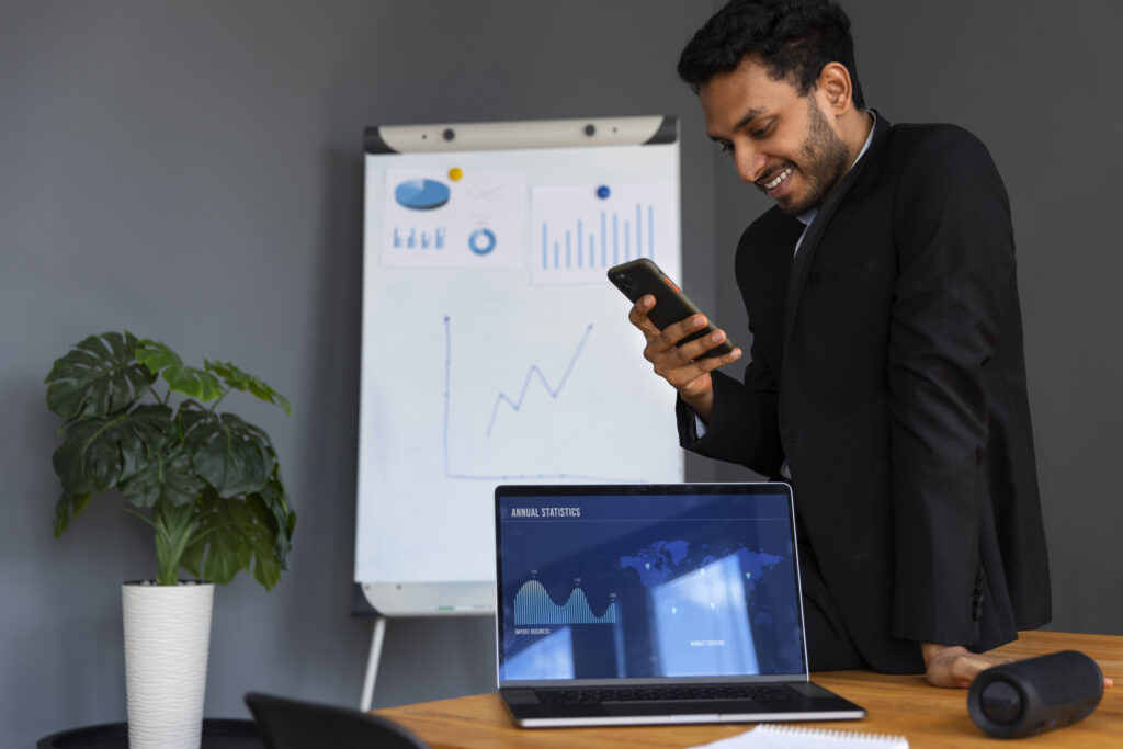 A business professional smiling while looking at his phone, with a laptop displaying annual statistics and a whiteboard with charts and graphs in the background.